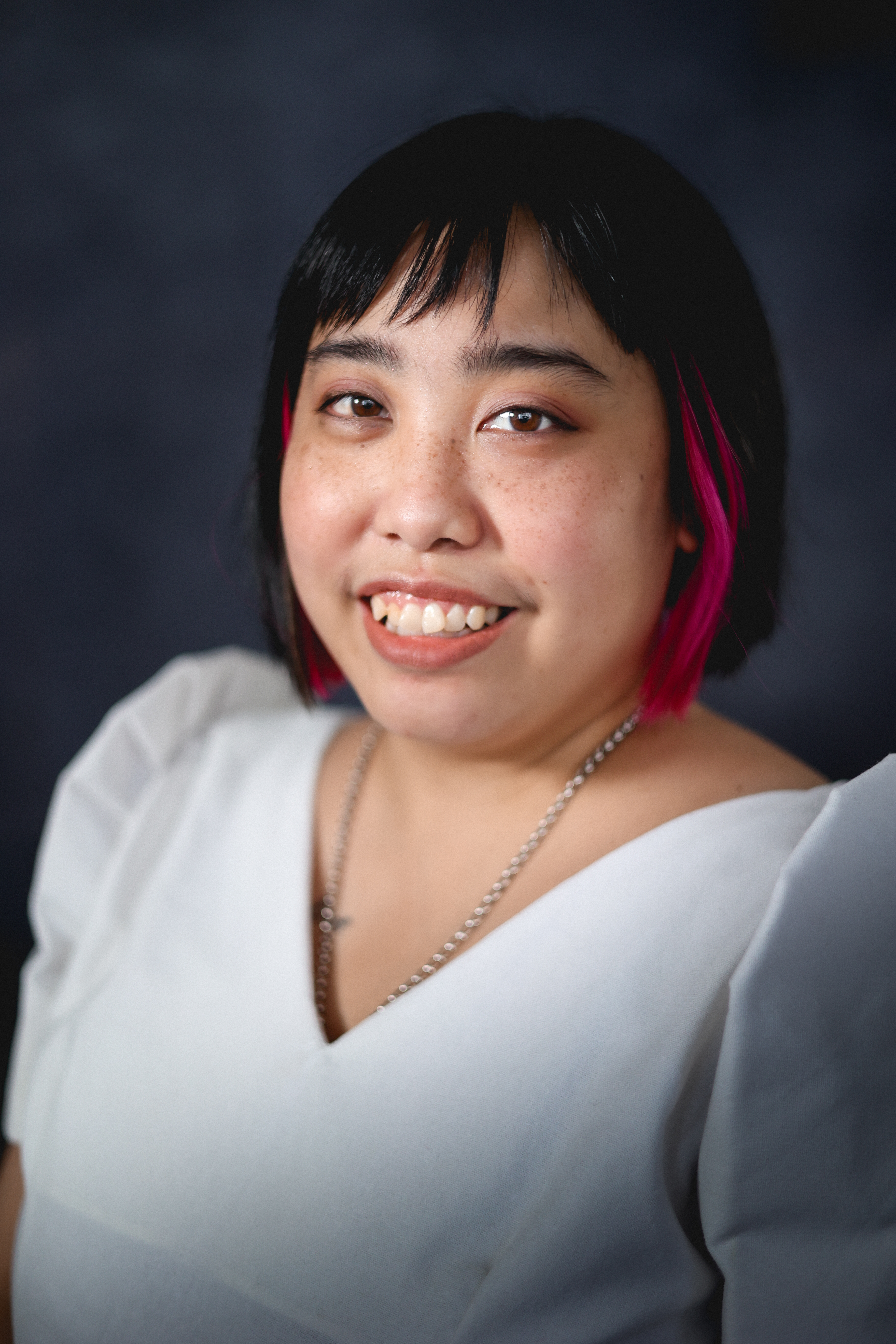 Caryl Loria Illana artist bio profile photo Caryl sits for a professional portrait. Caryl is Filipina. She's smiling broadly, her black hair with pink money piece streaks is styled in a sleek bob.